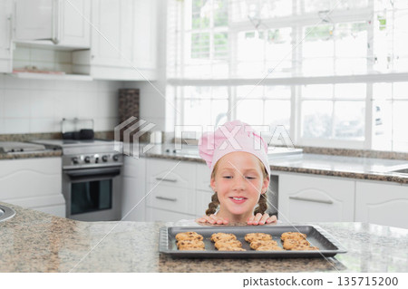 Girl wearing pink chef hat leaning on granite countertop in home kitchen, gazing at cookie tray Girl wearing pink chef hat leaning on granite countertop in home kitchen, gazing at cookie tray 135715200