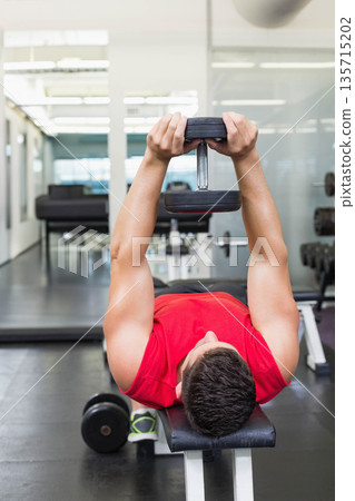 Lying male exerciser in red athletic top on bench in gym holding dumbbell by dumbbell rack Lying male exerciser in red athletic top on bench in gym holding dumbbell by dumbbell rack 135715202