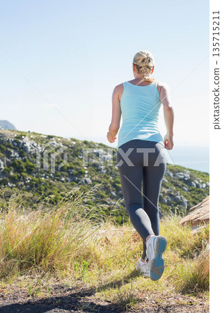 Adult woman jogging uphill on grassy coastal trail wearing light blue tank top gray leggings 135715211