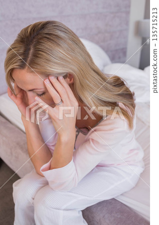 Woman pressing fingertips against temples and bowing head on bed edge at home wearing pink pajamas 135715213