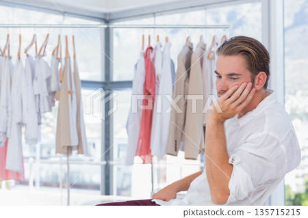 Man wearing white shirt sitting on stool in clothing showroom examining shirts on rack, copy space 135715215