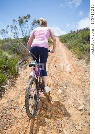 Woman riding purple bike with white water bottle, pedaling uphill on hillside trail in sportswear 135715216