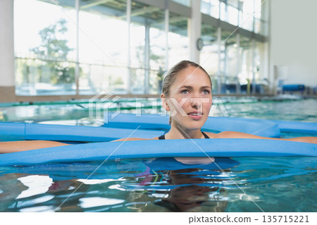 Adult woman wearing dark swimsuit leaning on blue foam flotation mats at pool under bright windows Adult woman wearing dark swimsuit leaning on blue foam flotation mats at pool under bright windows 135715221