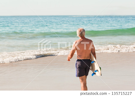 Mature male surfer walking into shallow turquoise surf on beach carrying shortboard Mature male surfer walking into shallow turquoise surf on beach carrying shortboard 135715227