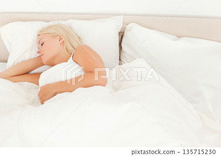 Female sleeping on bed with neutral headboard in bedroom wearing camisole with pillow and duvet 135715230