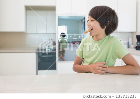Boy standing at kitchen island biting pizza slice wearing t-shirt beside built-in oven, copy space 135715235