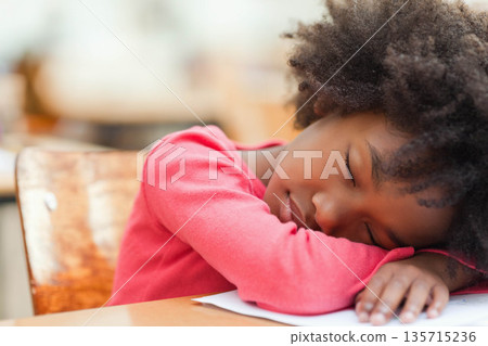 African American girl student resting head on folded arms on wooden desk with paper in classroom African American girl student resting head on folded arms on wooden desk with paper in classroom 135715236