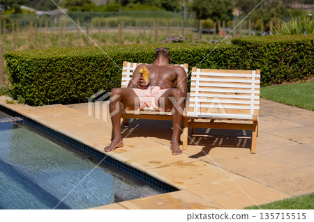 Reclining shirtless man in pink trunks sipping cocktail on bench beside pool, hedge visible 135715515