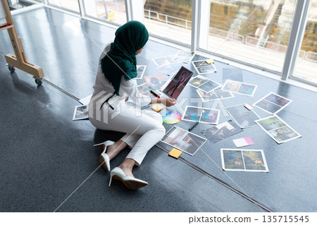 Middle Eastern hijabi woman sitting on studio floor, reviewing prints, color wheel and sticky notes 135715545