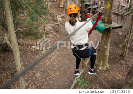 Female participant navigating ropes course in adventure park holding red lanyard, wearing helmet Female participant navigating ropes course in adventure park holding red lanyard, wearing helmet 135715550