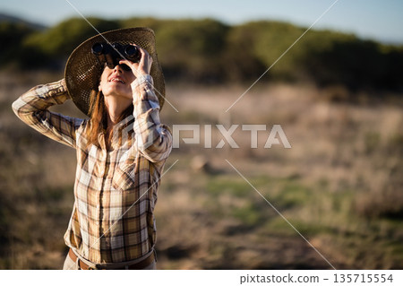 Woman standing holding binoculars and gazing upward in grassland wearing straw sun hat, copy space Woman standing holding binoculars and gazing upward in grassland wearing straw sun hat, copy space 135715554