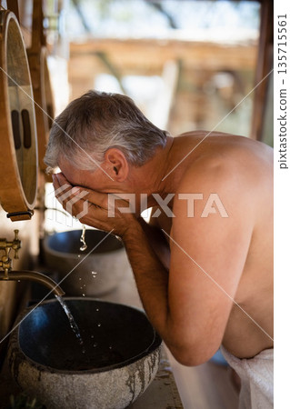 Senior man washing face over stone basin under brass faucet in open-air pavilion, copy space Senior man washing face over stone basin under brass faucet in open-air pavilion, copy space 135715561