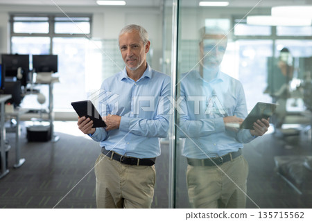Mature adult man leaning against glass partition and holding tablet at modern open-plan office Mature adult man leaning against glass partition and holding tablet at modern open-plan office 135715562