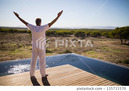 Middle-aged male stretching arms out on wooden pool deck by infinity pool overlooking scrub plain Middle-aged male stretching arms out on wooden pool deck by infinity pool overlooking scrub plain 135715570