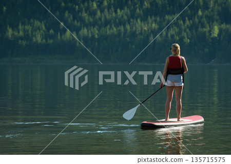 Female paddleboarder paddling on red paddleboard in lake wearing red vest, shorts, copy space 135715575