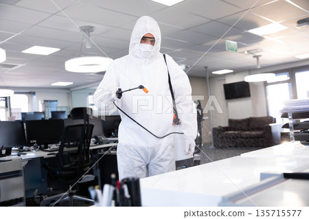 Man wearing white protective suit holding backpack sprayer wand disinfecting office desk area 135715577