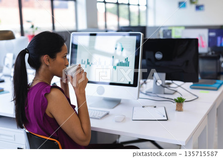 Senior Asian woman analyzing bar charts on monitors and holding coffee mug at office, copy space 135715587