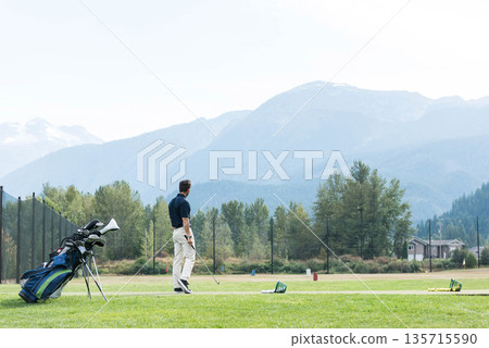 Male golfer practicing swing on tee mat at driving range with golf bag, copy space Male golfer practicing swing on tee mat at driving range with golf bag, copy space 135715590