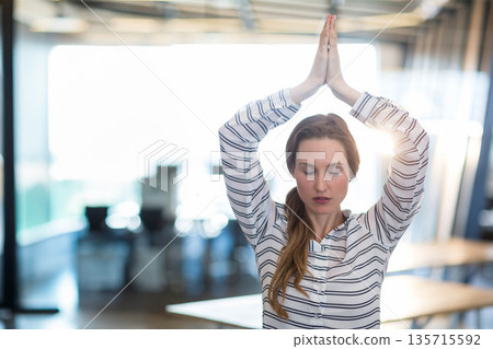 Woman wearing business attire holding yoga pose at desk in open-plan office with monitors Woman wearing business attire holding yoga pose at desk in open-plan office with monitors 135715592