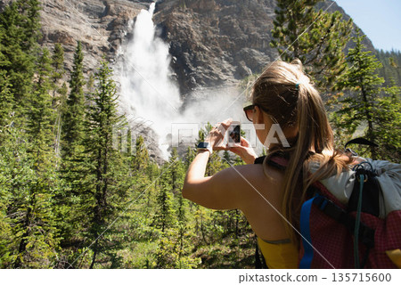 Mid adult female hiker with backpack and smartphone shooting waterfall on forest trail, copy space 135715600