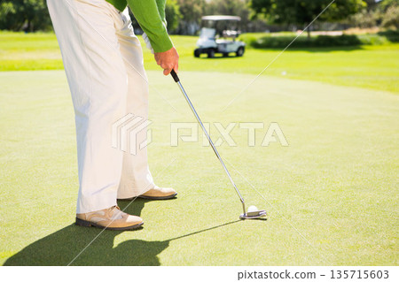 Senior African American man gripping putter and tapping golf ball on putting green near golf cart Senior African American man gripping putter and tapping golf ball on putting green near golf cart 135715603