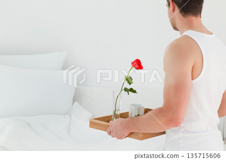 Man standing in bright bedroom holding wooden tray with rose and mug beside bed, copy space Man standing in bright bedroom holding wooden tray with rose and mug beside bed, copy space 135715606