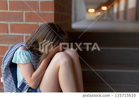Girl sitting on steps against brick wall hiding face with striped backpack at school, copy space 135715610