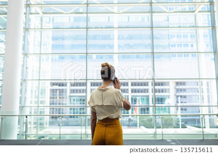 Woman holding smartphone standing at glass panels in terminal, with balustrade, skyline beyond Woman holding smartphone standing at glass panels in terminal, with balustrade, skyline beyond 135715611