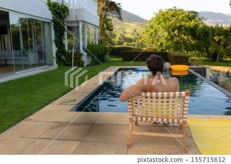 Shirtless middle-aged man sitting on woven lounge chair at house patio by pool with inflatable ring 135715612