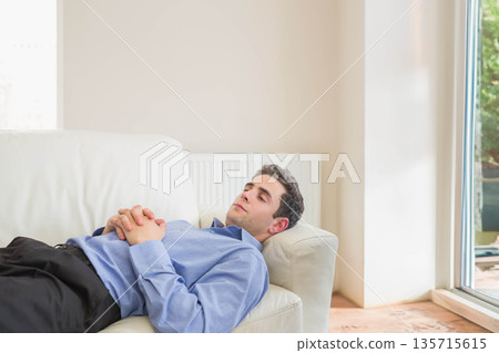 Man lying on light-colored leather sofa with cushion near radiator and large window in living room 135715615