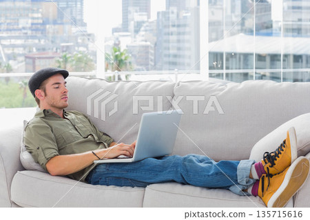 Man in his twenties reclining on light-gray sofa using silver laptop at home wearing yellow boots 135715616