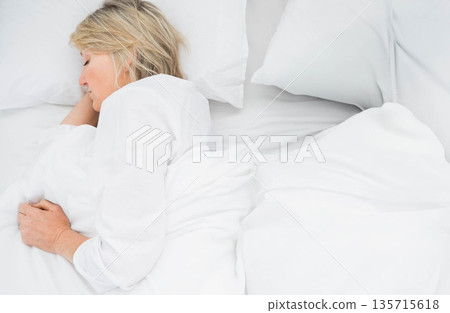 Middle-aged woman in white pajamas lying on left side on bed with white bedding, copy space 135715618