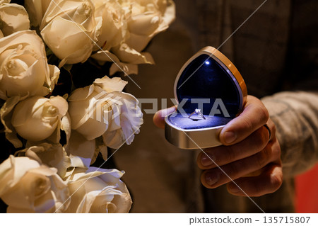 Heart-shaped ring box with diamond engagement ring illuminated by blue light beside white roses bouquet Heart-shaped ring box with diamond engagement ring illuminated by blue light beside white roses bouquet 135715807