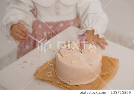 Young girl celebrating first birthday with pink cake and number one candle in soft pastel setting 135715809