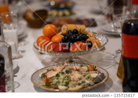 Elegant Table Setting with Fresh Fruit Platter and Caesar Salad in Glass Bowl on White Linen Tablecloth 135715817