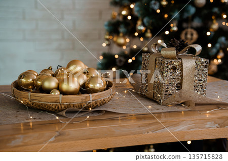 Golden Christmas Decorations and Gift Box on Wooden Table with Festive Lights and Tree Background Golden Christmas Decorations and Gift Box on Wooden Table with Festive Lights and Tree Background 135715828