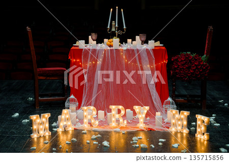 Romantic Proposal Setup with Illuminated "MARRY ME" Letters, Red Tablecloth, Candles, and Rose Bouquet on Stage Romantic Proposal Setup with Illuminated "MARRY ME" Letters, Red Tablecloth, Candles, and Rose Bouquet on Stage 135715856