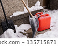 Snow-covered shovels and red fuel canister leaning against concrete steps during winter cleanup 135715871