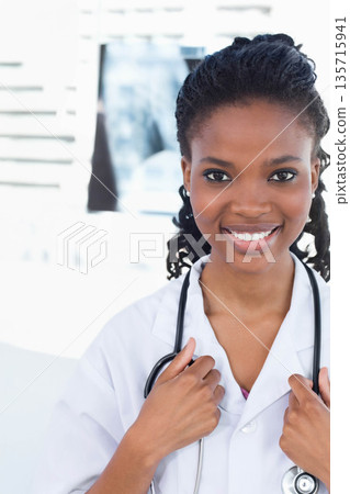 African American female clinician smiling, holding stethoscope and wearing white coat in clinic 135715941