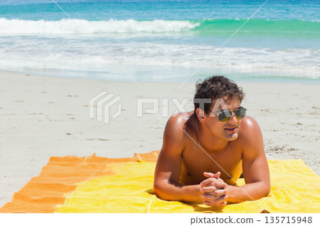 Man reclining on yellow-orange towel near shore wearing trunks and aviator sunglasses, copy space 135715948