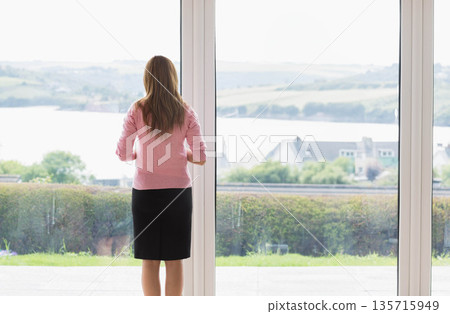 Woman standing at home in pink sweater, black skirt, gazing through glass doors at water 135715949