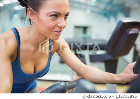 Woman working out on cardio machine at gym holding handles with blue sports tank top 135715950