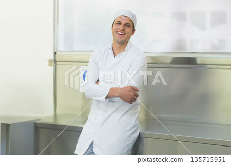 Mid-adult man in white chef coat and skullcap leaning at prep counter in kitchen smiling Mid-adult man in white chef coat and skullcap leaning at prep counter in kitchen smiling 135715951