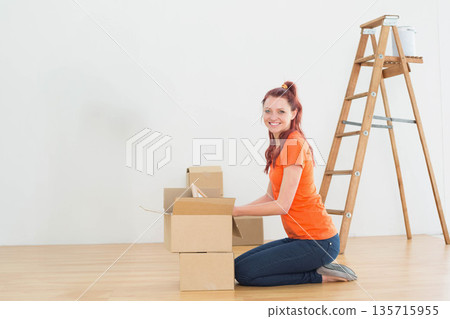 Female kneeling packing boxes on light floor in room near stepladder with paint bucket, copy space Female kneeling packing boxes on light floor in room near stepladder with paint bucket, copy space 135715955