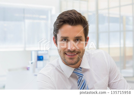 Man sitting at desk in modern office wearing white shirt and blue-striped tie, facing camera 135715958