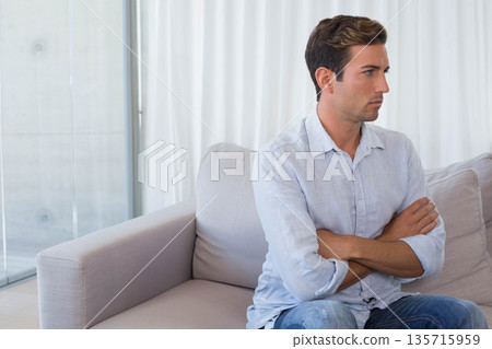 Man sitting on light gray sofa in living room, crossing arms and wearing blue shirt, jeans Man sitting on light gray sofa in living room, crossing arms and wearing blue shirt, jeans 135715959