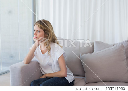 Adult woman resting chin on hand, sitting on gray sofa, wearing white top and dark jeans Adult woman resting chin on hand, sitting on gray sofa, wearing white top and dark jeans 135715960