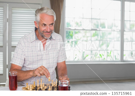Senior man wearing checked shirt playing chess on wooden chessboard at table with two tall glasses 135715961