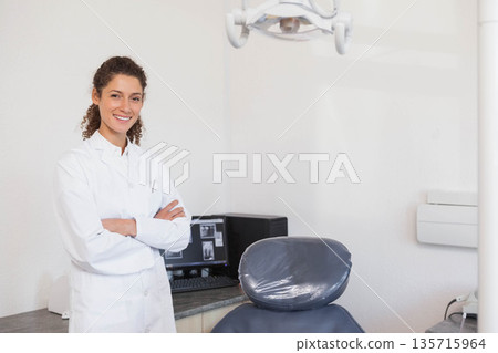 Female clinician wearing lab coat standing with arms crossed by chair covered plastic, copy space 135715964
