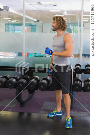 Adult man standing beside dumbbell rack holding water bottle in gym wearing blue gloves, copy space 135715966